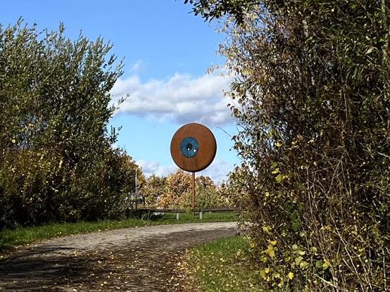 An der Biegung eines Weges, der von hohen Büschen gesäumt wird, steht vor einem blauen Himmel eine Skulptur, die einem Auge ähnelt.
