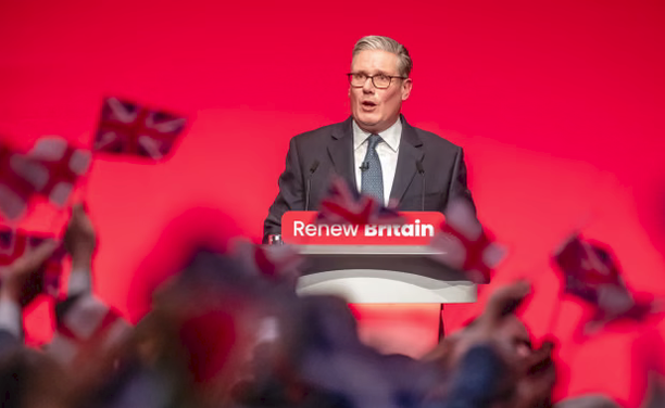 A politician stands in front of a red featuredless background wearing a dark suit and blue tie,  he has a sign on his podium that is red with white letter: "Renew Britain" and in front of him people are waving little union jacks.