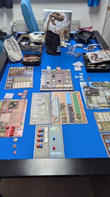 Tisch mit einem Brettspiel. Mehrere gemeinsame  Spielbereiche in der Mitte, zwei Spielertableaus, Münzen und andere Spielmaterialien sind zu sehen sowie der Spielekarton und die Anleitung.