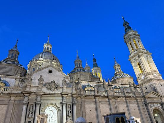 A historic cathedral with ornate domes and towers, illuminated against a deep blue evening sky. The facade features intricate sculptures and detailed architectural elements.
