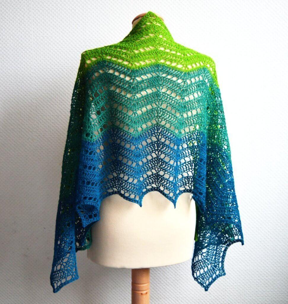 A rectangular crochet wrap, in a lacy chevron pattern and worked in light green, dark green and teal yarn. It's shown from the back on a mannequin against a white background.
