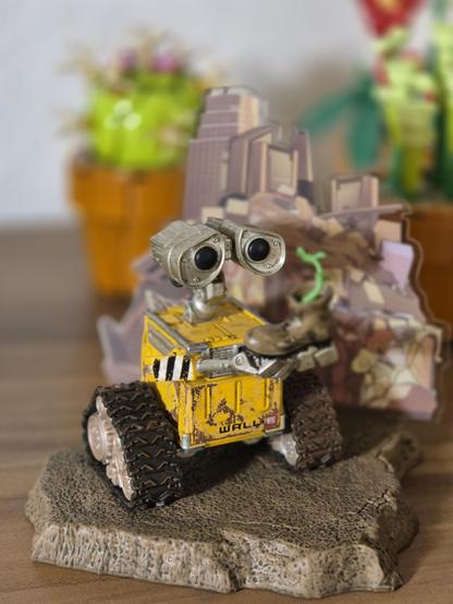 Perched atop a rugged, textured stone base is an endearing miniature figure of WALL-E, the beloved robot from the animated film. His bright, expressive eyes—each a perfect circle of curious wonder—sit atop a weathered yellow body, giving him an instantly recognizable charm. The details are delightful: his segmented treads, sturdy and ready for adventure, contrast with the delicate green sprout he cradles in his claw-like hand, a tender reminder of hope and renewal.

Behind him, a soft blur of v…
