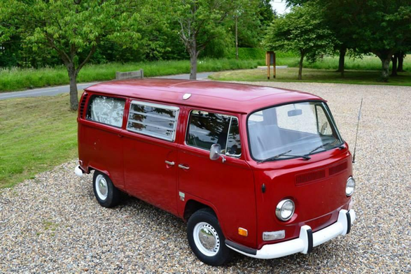 Red vintage VW bus (Volkswagen Type 2 van) parked on gravel, surrounded by green grass and trees in a peaceful outdoor setting.