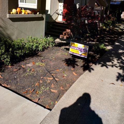 Small front yard of dirt with leaves in front of a green stucco wall with autumn pumpkins in a window ledge and a “Support School Care” yard sign near a shaded sidewalk.