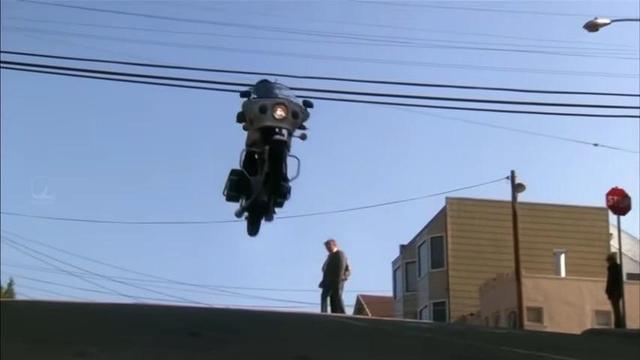 A motorcycle is airborne, jumping over a set of power lines, while a man stands on the ground watching. The scene is set against a clear blue sky.