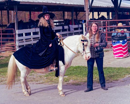 A woman with long blonde hair is wearing a witch hat and wearing a black dress riding a small cleam-colored horse with a woman wearing blue jeans, flannel shirt and blue jacket with cattle pens in the background and an American flag trashcan