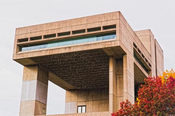 A concrete building has a large cantilevered section that juts out from a tower in the back and is supported on two legs,  one of which has a big notch visible from the front.  At the bottom a door opens onto a patio,  the cantilevered part has two layers of window,  a big wide one at the bottom and a set of subdivided smaller ones above and the bottom of the cantilevered section is covered by a grid that has numerous lights with a shimmering pattern