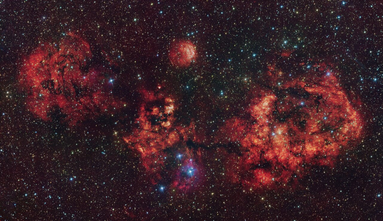 A telescopic view of a cosmic cloud against a dark starry sky. The cloud resembles a flying bat, spanning its wings. The cloud is mostly red, with some areas partially blocked by dark dust clumps.