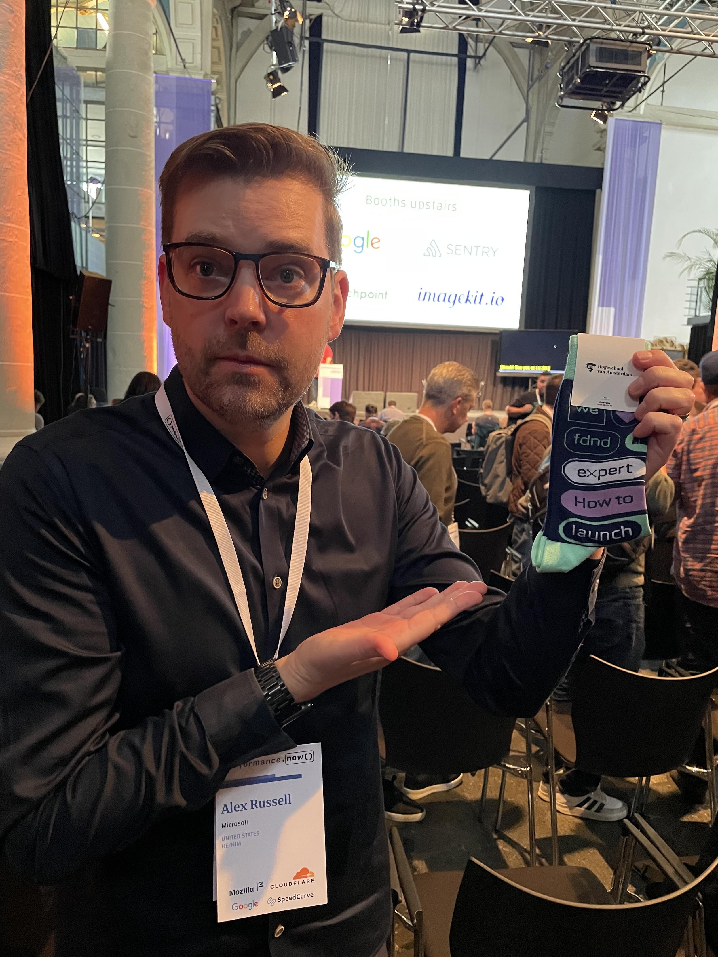 A man with glasses, holding a pair of socks, pointing towards it. The socks have terms like “fdnd”, “expert”, “how to” and “launch”, presented as separate pills in blue, white, turquoise and purple. A paper label wrapping the socks says “Hogeschool van Amsterdam”. The man wears a lanyard and performance.now() branded badge, saying “Alex Russell, Microsoft, United States, he/him”. In the background a conference venue with some chairs, a huge screen and some people are visible.