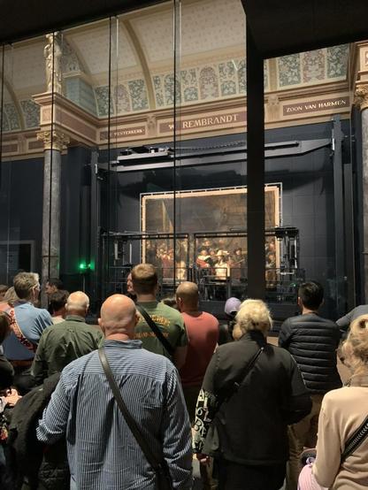 De Nachtwacht van Rembrandt van Rijn (figuren van een schutterscompagnie met sjerpen) op de achtergrond, op de voorgrond op de rug gezien bezoekers met schuine banden van tassen over hun rug.