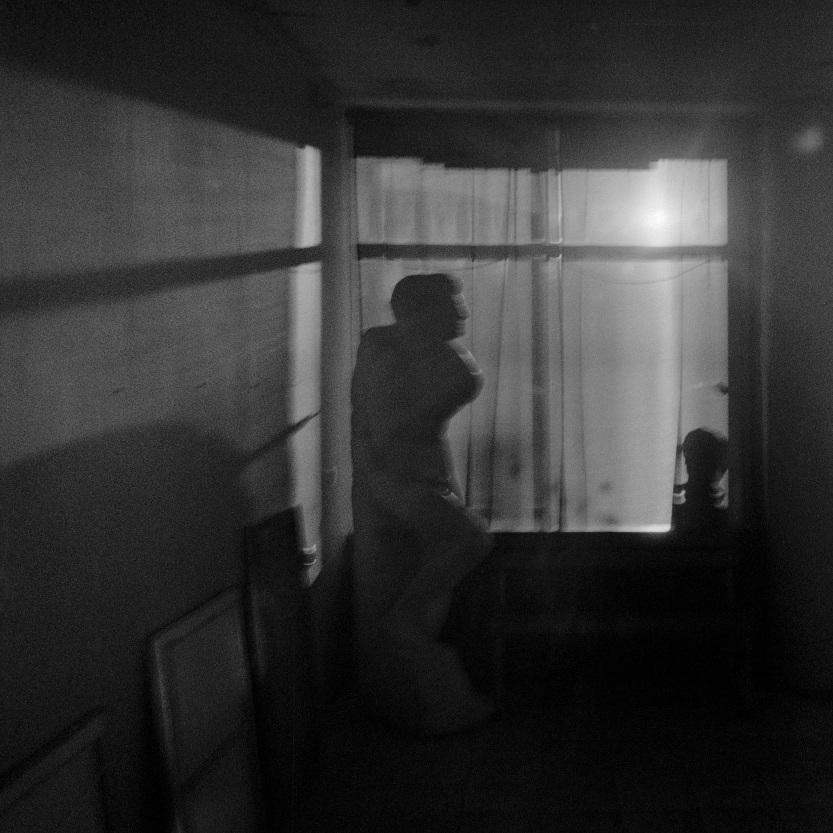 A black-and-white photo of an art school corner. Bright light falls from the window. Shadows are visible on the wall to the left. The paintings are located against the wall in the shade. A statue is placed near the window, and a bust silhouette is on the windowsill.