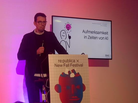 Dirk von Gehlen steht auf einer Bühne vor einem Bildschirm. Der Mann trägt ein schwarzes Hemd und eine Brille. Hinter ihm befindet sich ein Bildschirm mit Text und einem Cartoonbild. Vor der Bühne befindet sich ein Podest mit einem roten Cartoonbild eines Affenkopfes.

Der Bildschirm zeigt den Text „Aufmerksamkeit in Zeiten von KI“. Unterhalb des Textes befindet sich das Logo „re:publica x New Fall Festival“. In der oberen rechten Ecke des Bildschirms befindet sich ein kleines Logo mit dem Text…