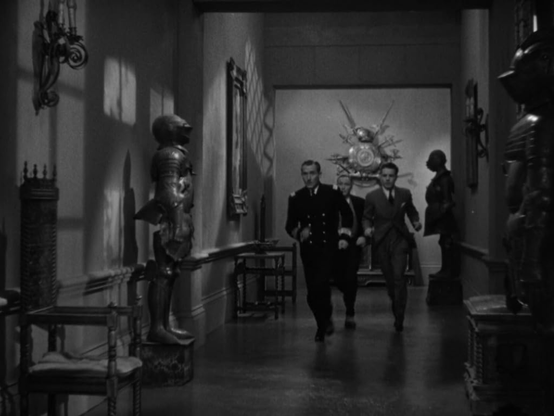 an image from the film of the men running quickly down a hallway, there is a suit of armor and axes and halberds on the walls of the old castle manor