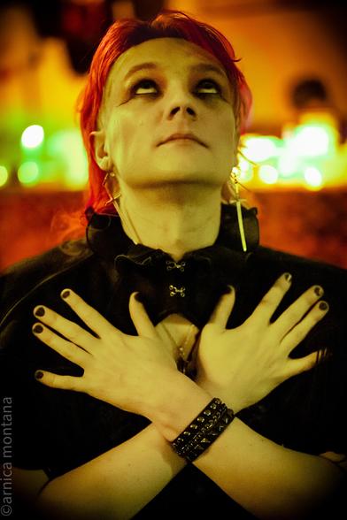 A person with bright red hair, wearing dark makeup and gothic attire, poses with arms crossed against their chest. The background features warm, colorful lights, creating an atmospheric effect.