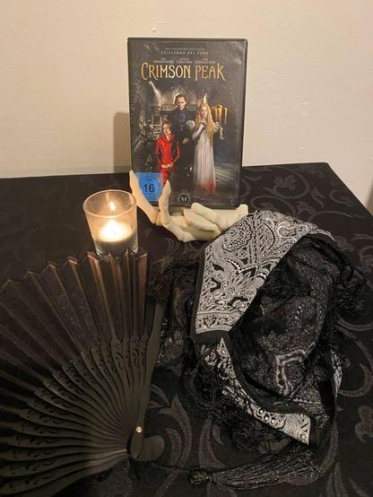 Photo: A "skeleton hand" holder is holding the DVD of Crimson Peak. There is a decorative black and white scarf with a nice pattern, and a black fan, also a burning tea light in a glass jar. All of it is placed on a table with a black table cloth.