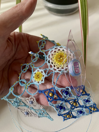 An elaborate unfinished project in tatting and beading, with harlequin motifs and beaded round elements, shown on a hand with a tatting shuttle ready to do a join. Colors are various shades of blue with gold and bright yellow.