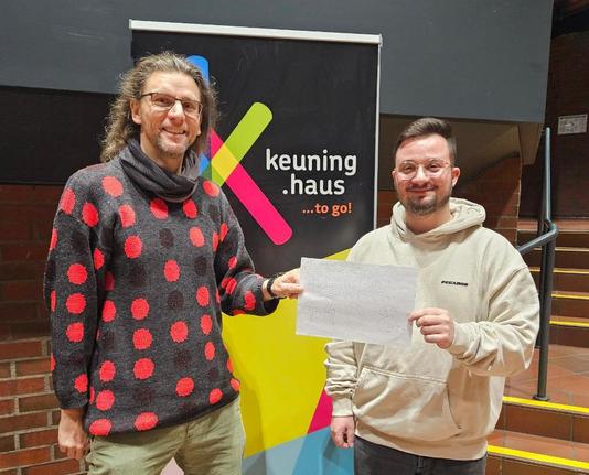 Auf dem Foto stehen zwei Männer nebeneinander und lächeln in die Kamera. Links steht Benjamin Beckmann mit langen, lockigen Haaren, Brille und einem dunkelgrauen Pullover mit großen roten Punkten. Rechts daneben steht Dominic De Marco mit kurzer Frisur, Brille und hellbeigem Hoodie. Beide halten gemeinsam einen handgemalten, symbolischen Spendenzettel im Format DIN A4 in die Kamera.
Hinter ihnen hängt ein großes, farbiges Banner mit dem Schriftzug „keuning.haus …to go!“.
Der Hintergrund zeigt Backsteinwände und eine Treppe im Inneren des Dietrich-Keuning-Hauses.