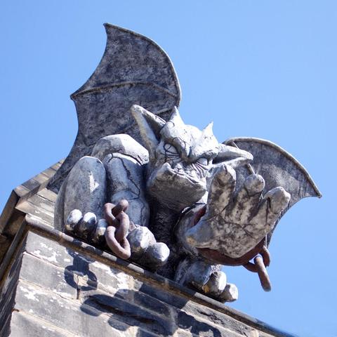 A close-up view of a stone gargoyle perched on the edge of a building, its wings raised and one clawed hand extended forward. The weathered statue has a fierce expression and is partially wrapped in a rusty chain. The background shows a clear blue sky.
