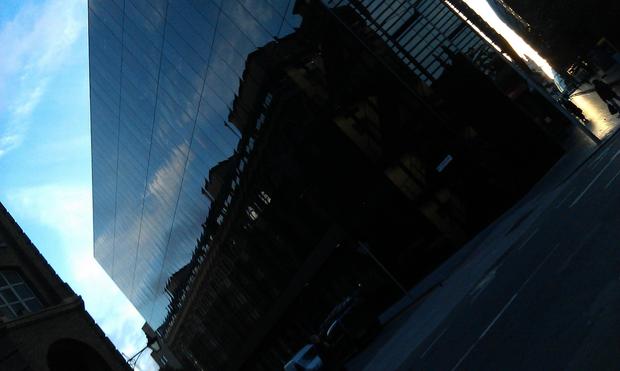 The facade of a slab of large glass building reflecting an adjacent large  building merges into a split of the light blue morning sky above.