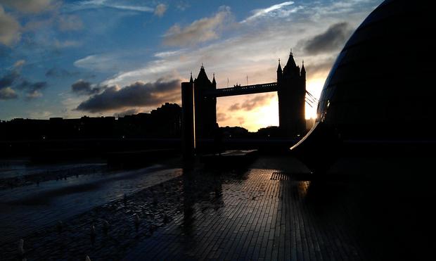 A silhouette of a landmark tower bridge almost frames a fierily rising sun which merges into a blue sky filled with  a variation of clouds dark to plain white in hue. The tower bridge flanked by more silhouettes of buildings and foliage, lines the horizon revealing a rain swept floor littered with fallen leaves towards the cameraman.