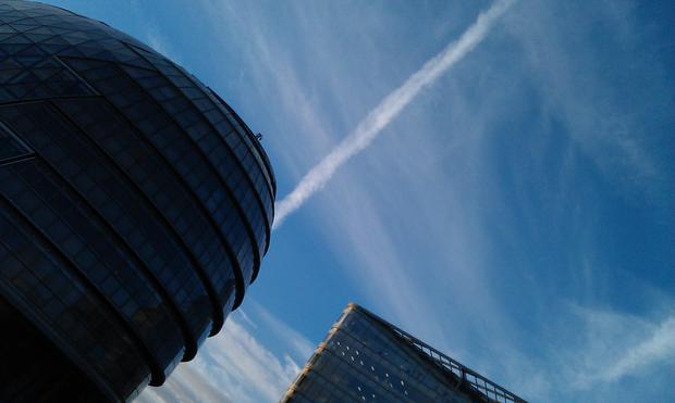 A gherkin shaped building almost in silhouette is partly visible captured at a 45 degree angle next to the edge of another block of offices against a blue but slightly clouded sky. A line of  dissolving smoke left by an aircraft seemingly emanates from the top side of the gherkin creating an interesting illusion.