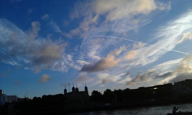 Silhouettes of city buildings and landmarks merge into a line of shapes splitting the horizon between an early morning detailed blue sky full of clouds  varied in hue, a dark line of river and  vast area of land.
