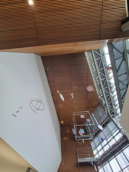 Daytime interior photo of sculpture art "Fertile Ground" by Cork Marcheschi. Various electic items hang from the ceiling in various geometric shapes and objects (e.g., eyeglasses, plates, wire-frame cubes, rectangles). This camera angle shows a bit more of the sculpture, including what looks like a metal orrery (i.e., a model of the planets around the sun), and a few stacked items which include a key.