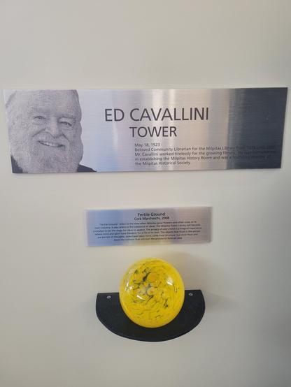 Daytime interior photo of two stainless-steel wall plaques, and a semi-circular shelf holding a blown-glass yellow orb, maybe several inches in diameter. The top plaque is larger, and shows the smiling face of an older bearded white male. The text reads "Ed Cavallini" and "Tower", and some tiny text about his contributions to the library. The bottom plaque is hard to read but it says "Fertile Ground" and "Cork Marcheschi, 2008", and talks a bit about how the art speaks to how libraries can represent catalysts for ideas to appear.