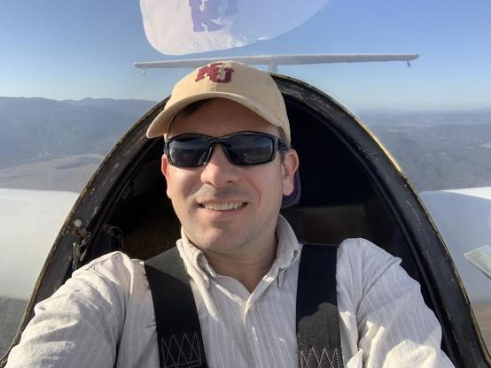 A smiling man in sunglasses and a hat sits in the cockpit of a glider in flight