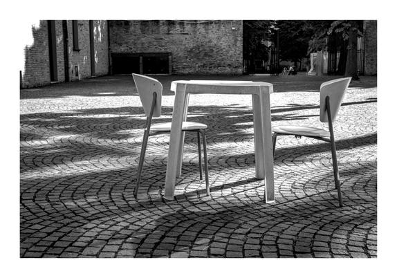 Zu sehen ist eine schwarz weiß Fotografie. In der Bildmitte steht ein Tisch mit zwei Stühlen. Die Gruppe steht auf gepflastertem Boden, welche in Bögen angeordnet ein Muster ergibt.