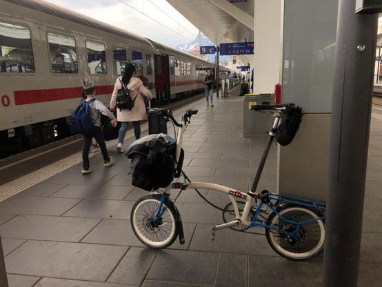 Ein Brompton Faltrad mit dicker Gepäcktasche steht an einem Bahnsteig, an dem gerade ein InterCity mit offenen Türen steht. Eine Frau und ein Kind mit Rucksäcken sind von hinten zu sehen, sie gehen den Bahnsteig entlang