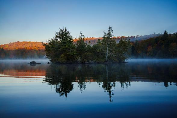 A small island with trees reflected in the still water of a lake, surrounded by morning mist and autumn colors.