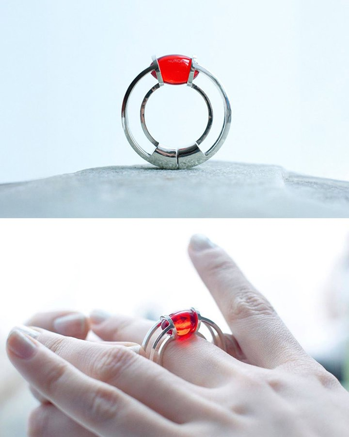 A two-panel photo showcasing a modern ring. The top panel shows the ring standing on a surface; it has a double silver band and a bright red gemstone. The bottom panel shows the ring being worn on a person's finger.