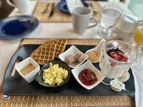 Frühstück.
Rechteckiger Teller mit vielen, kleinen Schälchen, zwei kleinen Waffeln und einem Glas Joghurt. In den Schälchen hat es Paté, Caramelcreme, Erdbeermarmelade, Rührei und Erdnussbutter. Saft und Kaffee daneben.
