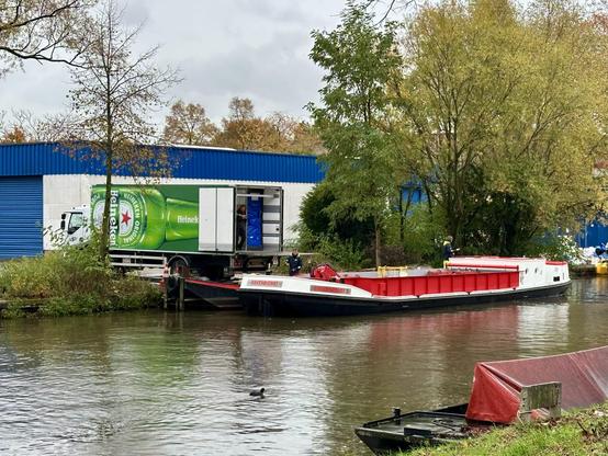 A dock area along a canal features a barge with a red cargo area and a truck displaying Heineken branding. A person is seen working on the barge.