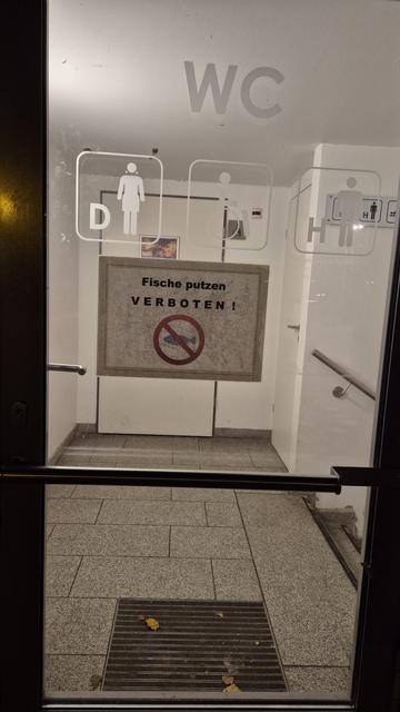 Der Eingang einer Toilettenanlage, der neben den üblichen Symbolen, wer hier sein Geschäft verrichten darf, auch folgendes Hinweisschild aufweist: Fische putzen verboten! Nebst durchgestrichenem Fisch.