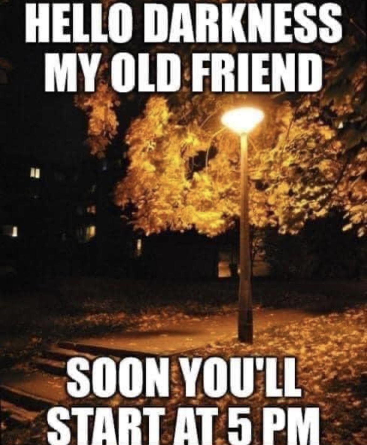 A streetlamp lit on a dark fall night. The caption is, “hello darkness my old friend. Soon you’ll start at 5pm”