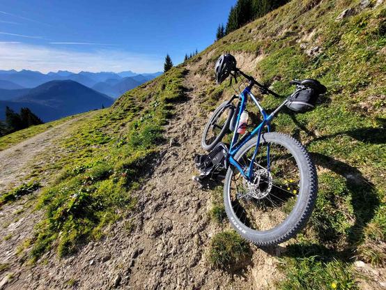 Perched on the edge of a rugged mountain trail, a sturdy blue mountain bike takes a well-deserved rest against the backdrop of an endless alpine panorama. The bike, equipped with wide, grippy tires and a sleek black frame bag, leans casually against the slope, its handlebars turned slightly as if gazing out at the view. The trail itself is a mix of sun-baked dirt and scattered rocks, winding through patches of vibrant green grass and wildflowers that dot the hillside.

Beyond the immediate fore…