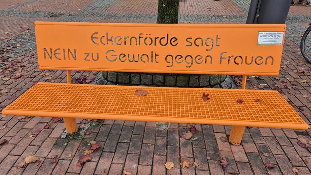 Eine orangefarbene Sitzbank aus Metal, umgeben von Herbstblättern, auf der zu lesen ist: Eckernförde sagt NEIN zu Gewalt gegen Frauen.