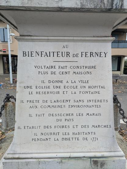 Texte sur la stèle de la statue "au bienfaiteur de Ferney. Voltaire fait construire plus de cent maisons. Il donne à la ville une église, une école, un hôpital, le réservoir et la fontaine. Il prête de l'argent sans intérêts aux communes environnantes. Il fait dessécher les marais du pays. Il établit des foires et des marchés. Il nourrit les habitants pendant la disette de 1771."