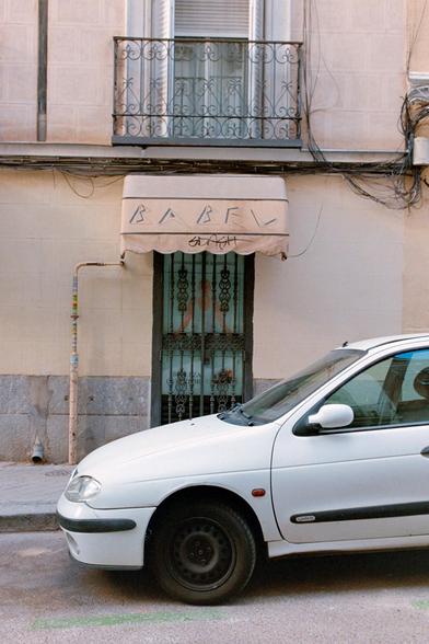 Photograph of a hair salon with an awning. The word "Babel" is written on it in a very 80s font. In the foreground, there is a car.

Fotografía de una peluquería con un toldo. En él se lee "Babel" con una tipografía muy ochentera. Delante, en primer plano, hay un coche.