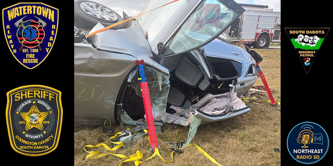 Crash scene with overturned car on grass, supported by stabilizers. Fire truck in background. Multiple agency logos on sides.