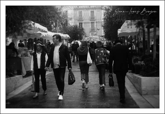 This black-and-white photograph captures a lively street scene filled with pedestrians moving in different directions. The monochrome tone lends a timeless, classic quality to the image, emphasizing contrasts of light and shadow.
In the foreground, two pairs of people walk side by side. On the left, a woman and a man appear engaged, perhaps in conversation, with the woman smiling gently. The man beside her has a casual yet confident stride, carrying a bag in his left hand. They stand out with their lighter clothing, especially the man's open jacket and white sneakers that pop against the cobblestones.
To the right, another pair strolls away from the camera. One wears a star-patterned jacket, which draws attention with its boldness even without color. The other is dressed more formally, possibly in black, suggesting a dynamic contrast not only in style but in mood or activity.
In the middle distance, a third individual—a woman—walks alone with a scarf wrapped around her neck. Her casual yet purposeful walk contrasts with the more social groups around her.
The background reveals an urban setting with trees and traditional architecture, characteristic of European city streets, lined with vendor stalls or market booths under small canopies. This suggests a market or fair atmosphere, inviting community interaction and commerce.
