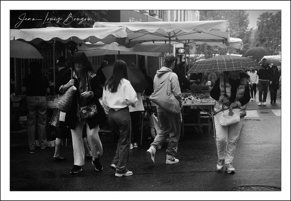 This black-and-white photograph captures a lively street scene, bustling with people seemingly engaged in casual, everyday activities. The choice of monochrome adds a timeless, almost nostalgic quality, enhancing the textures and contrasts in the image.
In the foreground, a group of pedestrians crosses the street, each person distinct in posture and movement. One woman, draped in a light jacket, confidently strides forward while a man beside her leaps playfully, adding a dynamic rhythm to the scene. Another woman, holding an umbrella, stands out against the pavement’s wet sheen, hinting at recent rain. The reflective surface of the road amplifies the glistening effect, lending an atmospheric detail to the photo.
The backdrop reveals a tented market stall, under a scalloped canopy, suggesting a community market or fair environment. People gather there, some seemingly engrossed in browsing or buying, adding layers of human interaction and commerce. The blurred figures further in the distance create depth and suggest an active, vibrant street life.
Visually, the contrast between the dark clothing of some individuals and the lighter apparel of others, combined with the shade variations, emphasizes movement and volume. The small details — like the person clutching a shopping bag, or the umbrellas held against an overcast sky — evoke a sense of everyday life and human resilience despite the rain.

