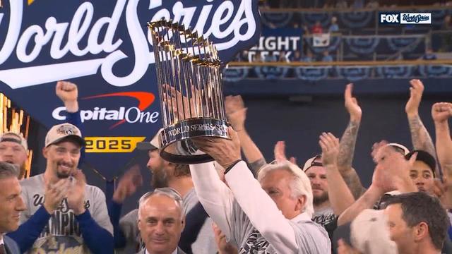 A member of the Los Angeles Dodgers organization proudly lifts the World Series championship trophy high above his head as teammates cheer and clap around him. Everyone is wearing gray “World Series Champions 2025” shirts and white caps. Behind them, a giant “World Series” logo with the Capital One sponsor glows on the big screen, surrounded by fireworks and celebration lights.