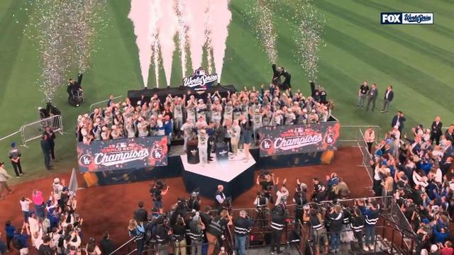 A wide overhead shot of the Dodgers’ 2025 World Series championship celebration shows the entire team gathered on a stage set up on the field. Confetti and fireworks burst into the air as players cheer and raise their arms. The stage banners read “2025 World Series Champions,” with media crews, photographers, and fans surrounding the scene and capturing the moment from all angles.