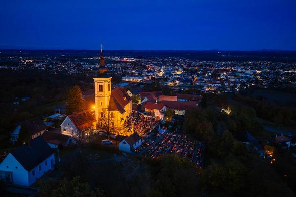 
Eine Drohnen Aufnahme bei tiefer Blauer Stunde zeigt die beleuchtete Wallfahrtskirche Frauenberg auf einem Hügel, umgeben vom Friedhof.

Die Kirche mit ihrem barocken, gelb beleuchteten Kirchturm und dem Hauptschiff dominiert das Zentrum. Die Dächer sind in warmem Rotbraun gehalten. Rechts von der Kirche und unterhalb im Hang sind zahlreiche Kerzen oder Lichter am Friedhof zu Allerheiligen zu sehen. Mehrere kleine Häuser mit weißen Fassaden und rotbraunen Dächern liegen in der Nähe.

Direkt hinter dem Hügel erstreckt sich die nächtliche Stadt Leibnitz. Die Gebäude und Straßen sind durch unzählige, größtenteils gelb-orangefarbene Lichter erhellt, die ein dichtes, warmes Netz über die dunkle Landschaft legen.

Ein tiefblau-dunkler Nachthimmel spannt sich über das gesamte Motiv und bildet einen starken Kontrast zu den warmen Lichtern.

Das Bild fängt die feierliche und stille Atmosphäre eines Gedenkabends ein.