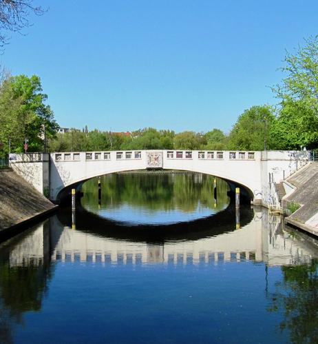 Eine weiße Brücke aus Stein überspannt einen Fluss. Sie spiegelt sich horizontal im Wasser. 
Das Ufer ist befestigt. Die Brücke hat ein Geländer mit lauter quadratischen Geflechten aus Metall. In der Mitte ein Wappen. Links und rechts ist sie mit einem Brückenkopf versehen. Der Himmel ist blau und die Vegetation aus Bäumen frühlingshaft grün.