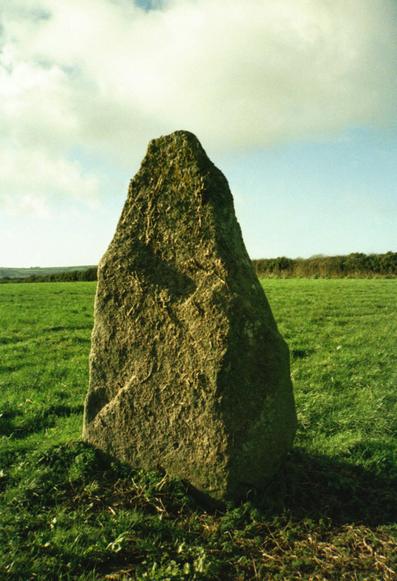 One of a pair of prehistoric standing stones or menhirs known as 'The Sisters', standing a short distance away from it's companion, within a field of short grass beneath a beautiful blue sky with many clouds. The stone is shaped a bit like an inverted canine tooth, The hedge-line curves gently upwards to the right in the middle distance. Behind the hedge, on the left, can just be seen the edge of the fields and moors beyond.