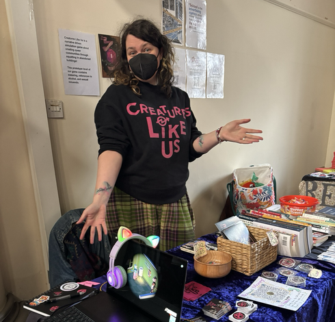 Tall white personal with curly hair and a Creatures Like Us sweater posing by a stall with a laptop, stickers and prints. 